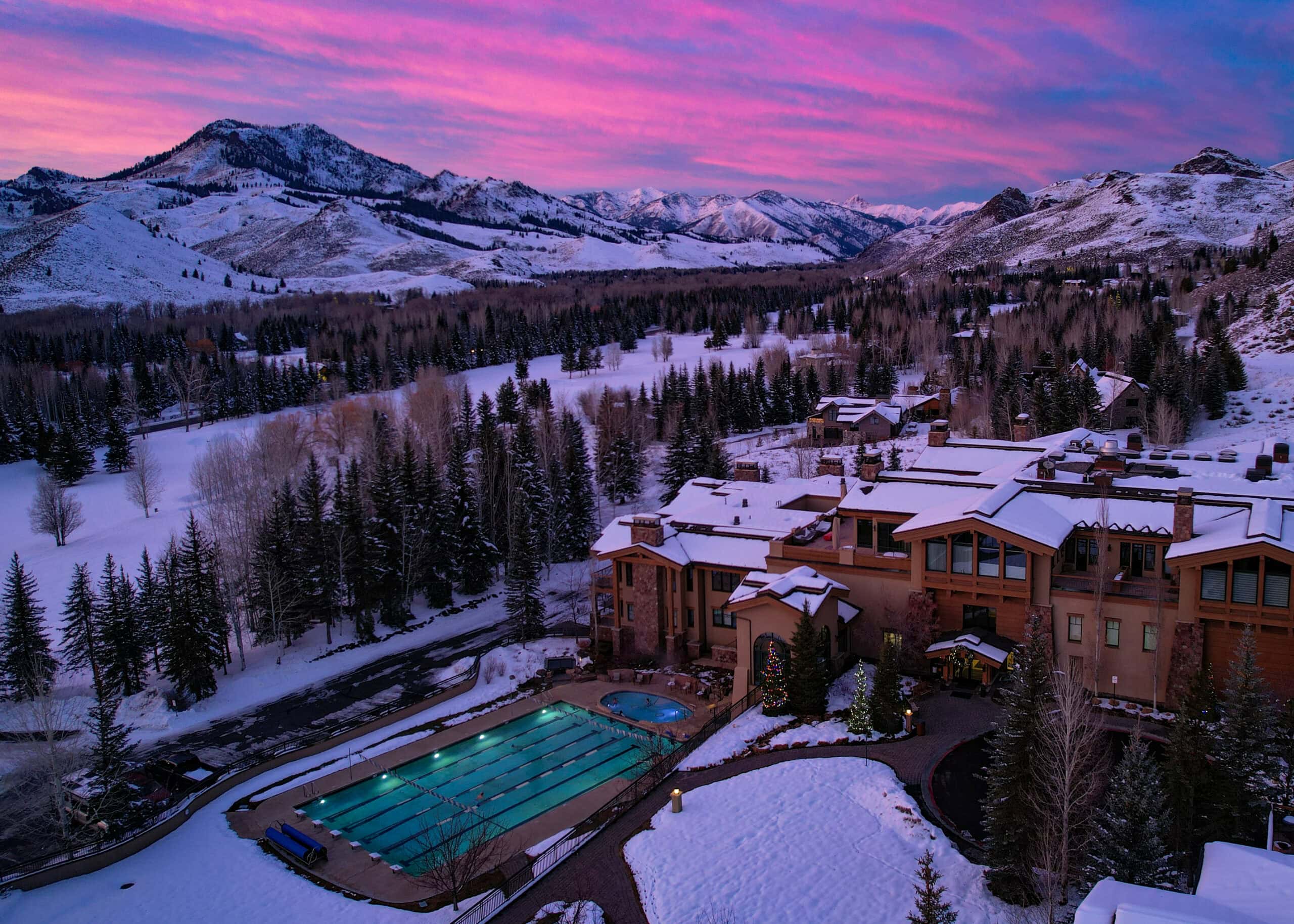 A large mountain lodge with snow-covered roofs sits beside a heated outdoor pool, surrounded by snowy trees and mountains under a vibrant pink and purple sunset sky. The inviting atmosphere evokes the tranquility of a Sun Valley spa, perfect for unwinding after a day of adventure in the crisp mountain air.