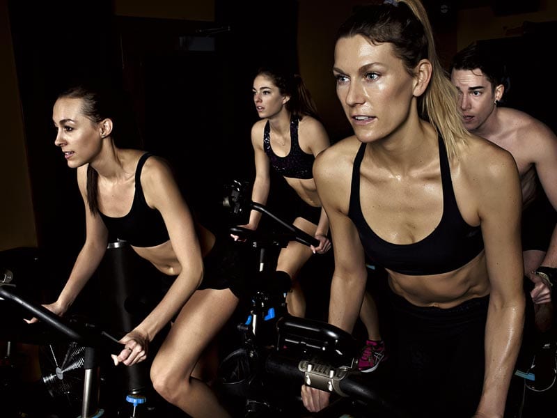 a man and three women ride to a sheen of sweat during an indoor cycling class
