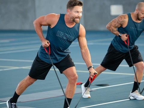 Two men exercise outdoors on a sports court using resistance bands, both wearing blue tank tops and black shorts, lunging forward with focused expressions. Their dedication to fitness reflects the vibrant wellness culture found at a Sun Valley health club, where strength training and holistic well-being go hand in hand.