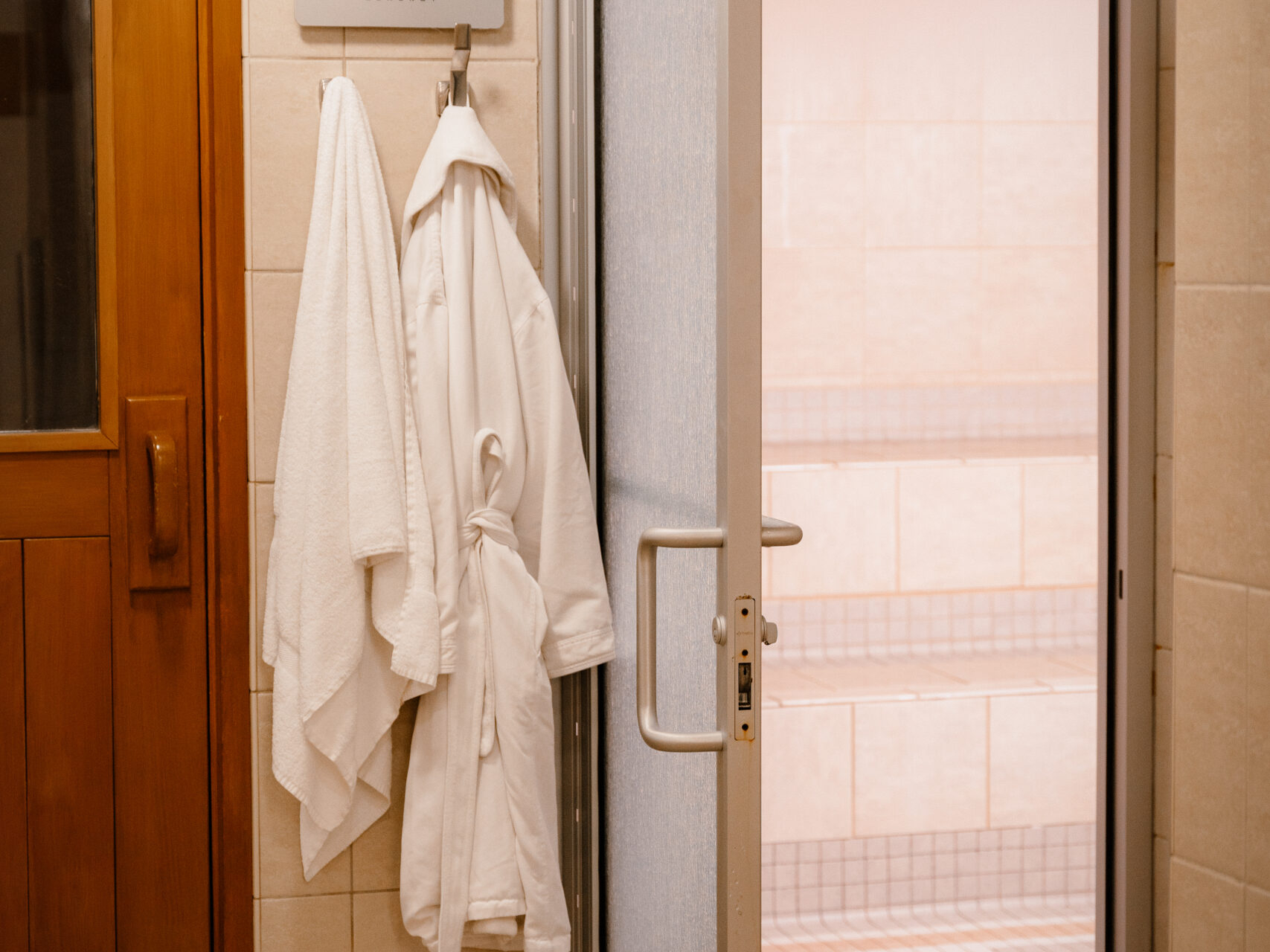 Two white bathrobes hang on hooks next to an open frosted glass door in a tiled Sun Valley spa or sauna area.