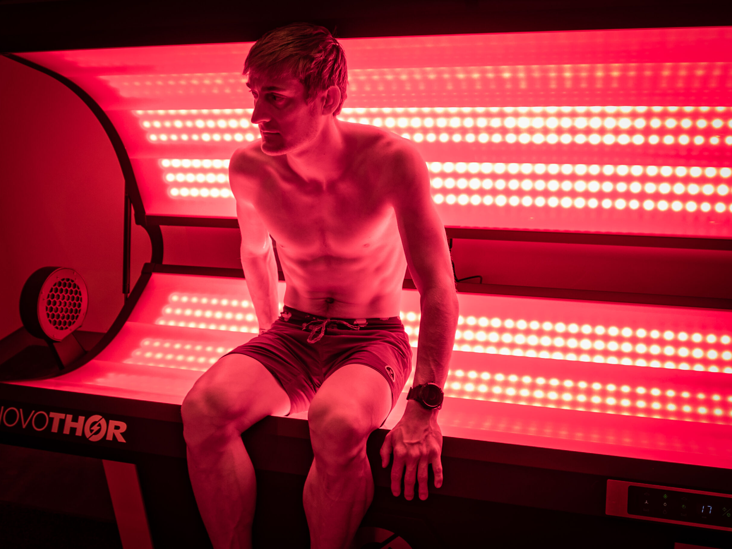 Shirtless person in shorts sits on the edge of a red light therapy bed, illuminated by bright LED panels inside a dimly lit room at a Sun Valley health club.