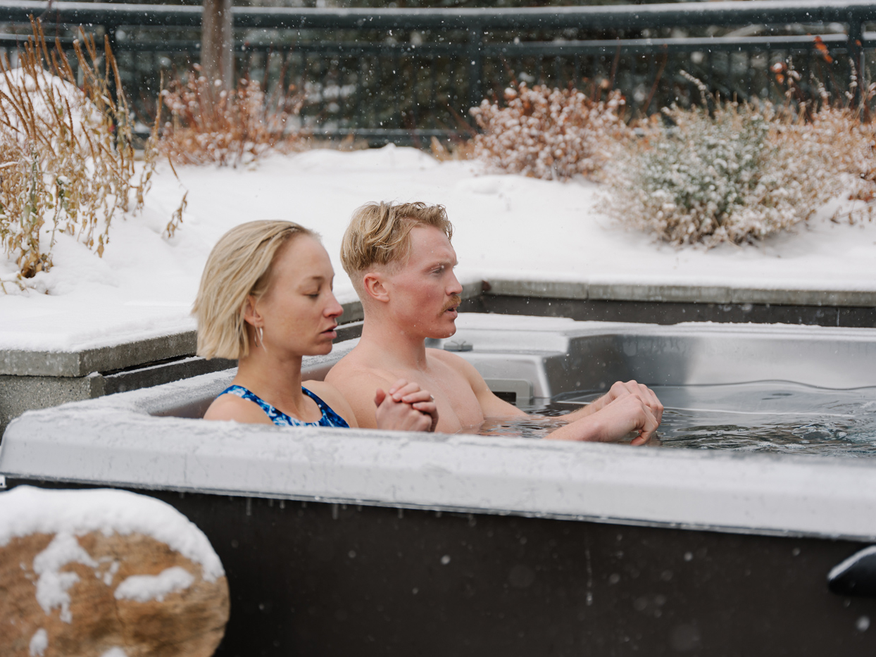 Two people sit side by side in an outdoor hot tub during winter, surrounded by snow and bare plants, enjoying a tranquil moment at the Sun Valley spa.