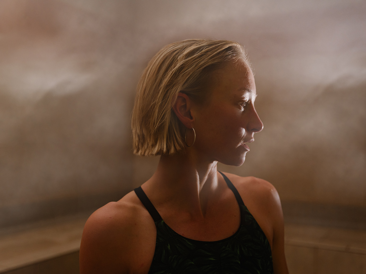 A person with short blond hair and hoop earrings stands in a steamy room at a Sun Valley health club, wearing a dark tank top, looking to the side with soft light highlighting their profile.