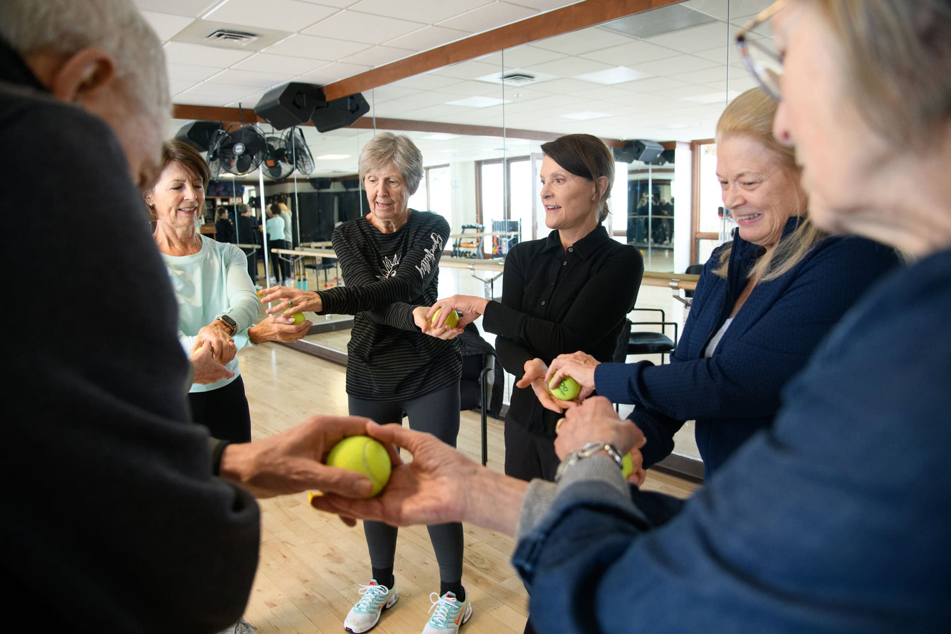 A group of older adults stands in a circle holding tennis balls and smiling in a brightly lit exercise room with mirrors at the Sun Valley health club.