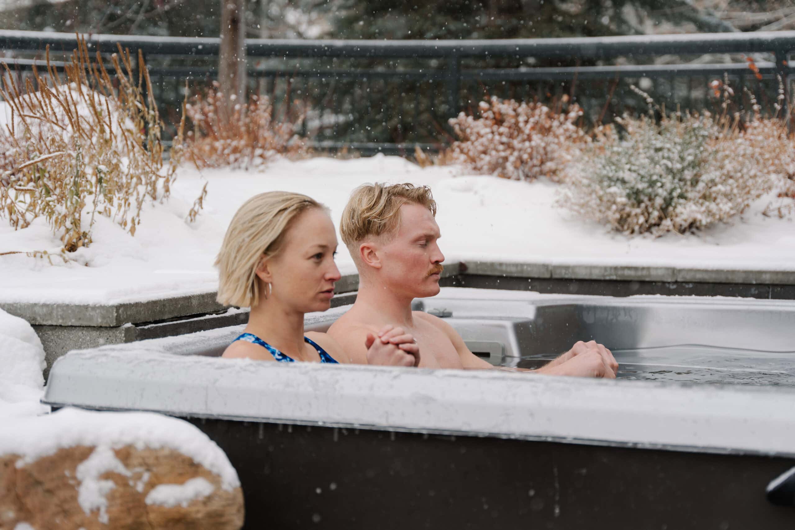 Two people sit side by side in an outdoor hot tub surrounded by snow-covered plants and trees, with snowflakes falling around them—a serene scene reminiscent of the tranquil experiences at a Sun Valley spa.