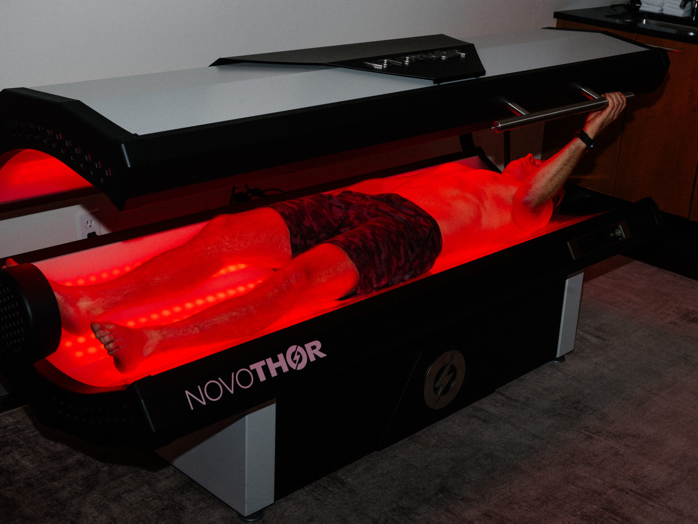 A person lies inside a NovoTHOR red light therapy bed at a Sun Valley spa, holding the lid open as the device emits vibrant red light onto their body in a dimly lit room.