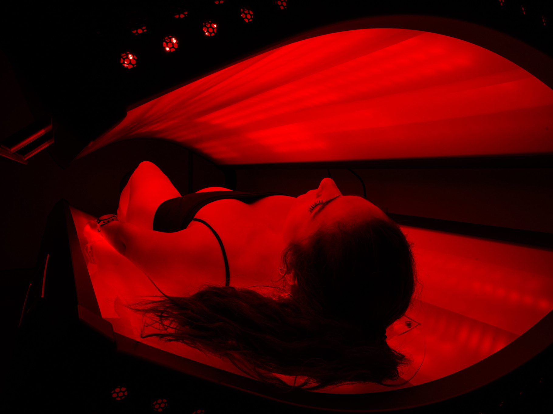A person in a bikini lies inside a tanning bed emitting red light at the luxurious Sun Valley spa, with their head resting back and long hair spread out.