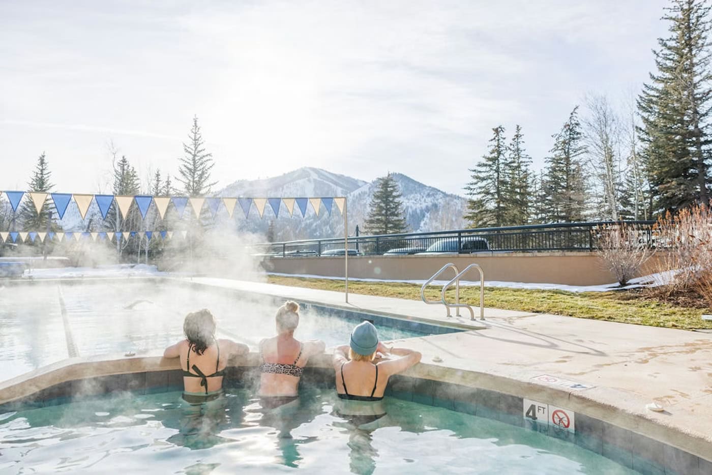 Three people in swimsuits relax in a steaming outdoor pool at a Sun Valley spa, surrounded by snowy mountains and pine trees on a sunny winter day.