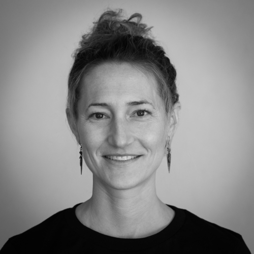 A woman with light hair in a bun, wearing earrings and a dark top, smiles at the camera against a plain background. The image is in black and white.