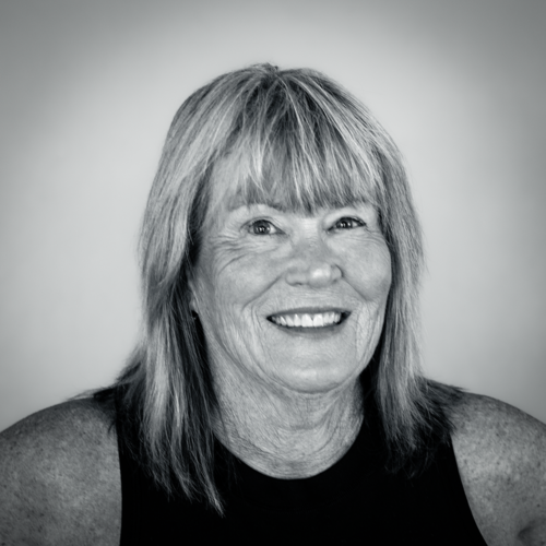 An older woman with shoulder-length light hair and a sleeveless black top smiles at the camera against a plain background. The image is in black and white, evoking the calm and timeless ambiance reminiscent of a relaxing Sun Valley spa.