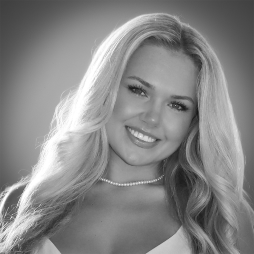 Smiling woman with long, wavy blonde hair wearing a pearl necklace and a light-colored top, photographed in black and white.