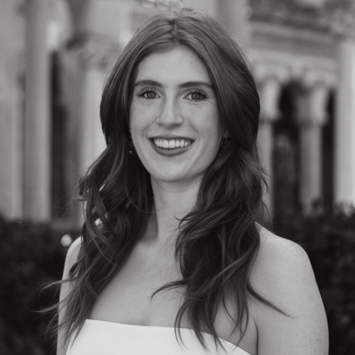Young woman with long wavy hair smiles at the camera, standing outdoors in front of a blurred building with arched windows. The image is in black and white.
