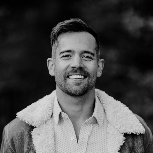 A man with short hair and a beard smiles at the camera, wearing a shearling-collared jacket and a patterned shirt, with a blurred outdoor background.