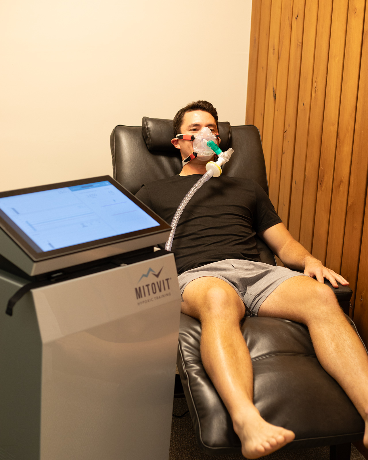 A man lies on a recliner wearing a breathing mask connected to a machine labeled “MITOVIT” in a room with wood-paneled walls, highlighting the Ultimate Recovery Destination where personal trainers oversee every step of your rejuvenation journey.