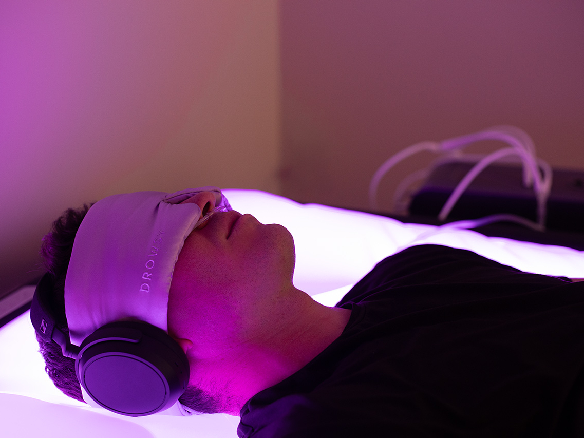 A person lies on a glowing surface, wearing headphones and an eye mask with “DRO” visible, in a dimly lit room with purple lighting—the Ultimate Recovery Destination designed to help you fully unwind.
