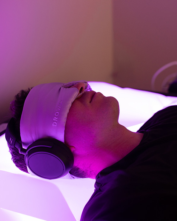 A person lies on a glowing bed wearing large headphones and an eye mask, illuminated by purple and white light, experiencing the Ultimate Recovery Destination.