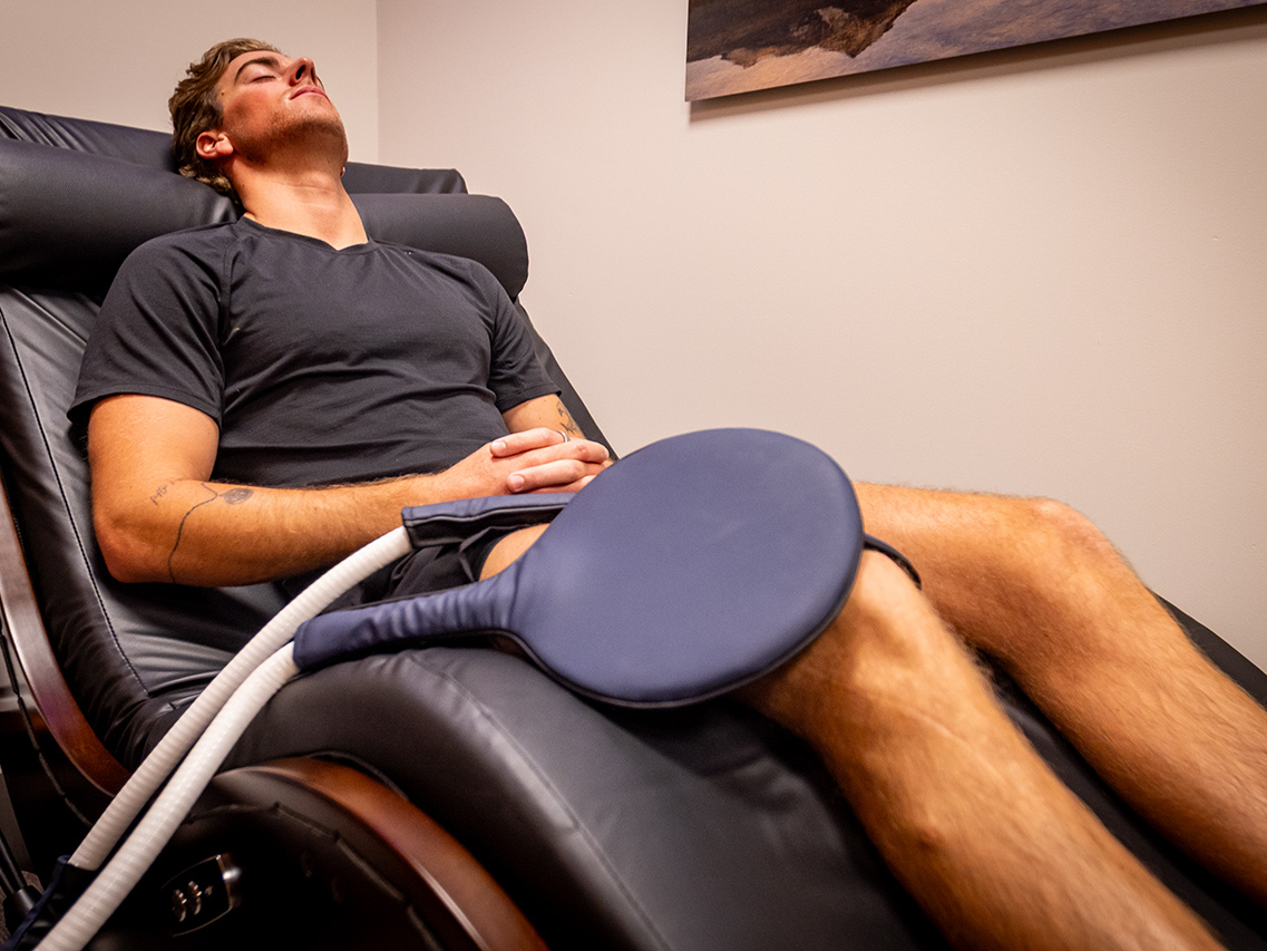 A person reclines in a chair with eyes closed, hands folded, and a round medical device resting on their thigh in a clinical setting—an experience that reflects the atmosphere of the Ultimate Recovery Destination trusted by personal trainers.