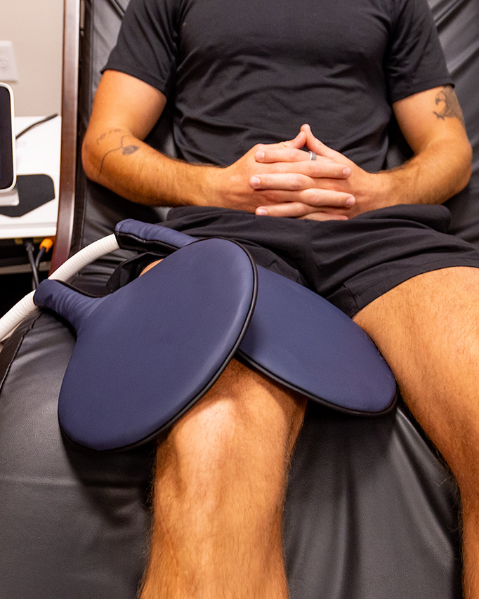 Person lying on a reclining chair with two large medical pads placed on their thighs; a medical device with display is on a nearby table—an essential scene at the Ultimate Recovery Destination, where personal trainers tailor every aspect of your wellness journey.