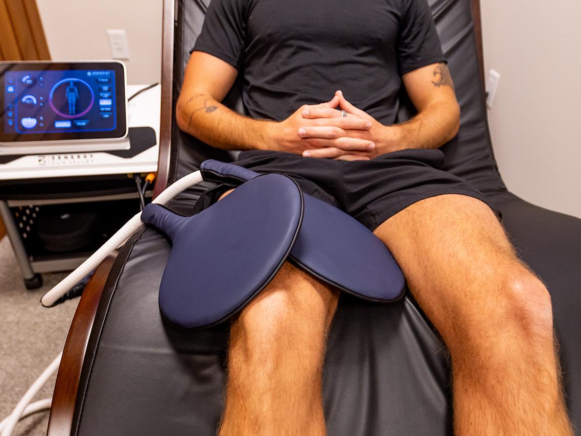 A person sits on a reclined chair with two round pads placed on their thighs, enjoying the benefits at the Ultimate Recovery Destination. A medical device with a display screen is visible nearby, creating an environment trusted by personal trainers for advanced recovery techniques.