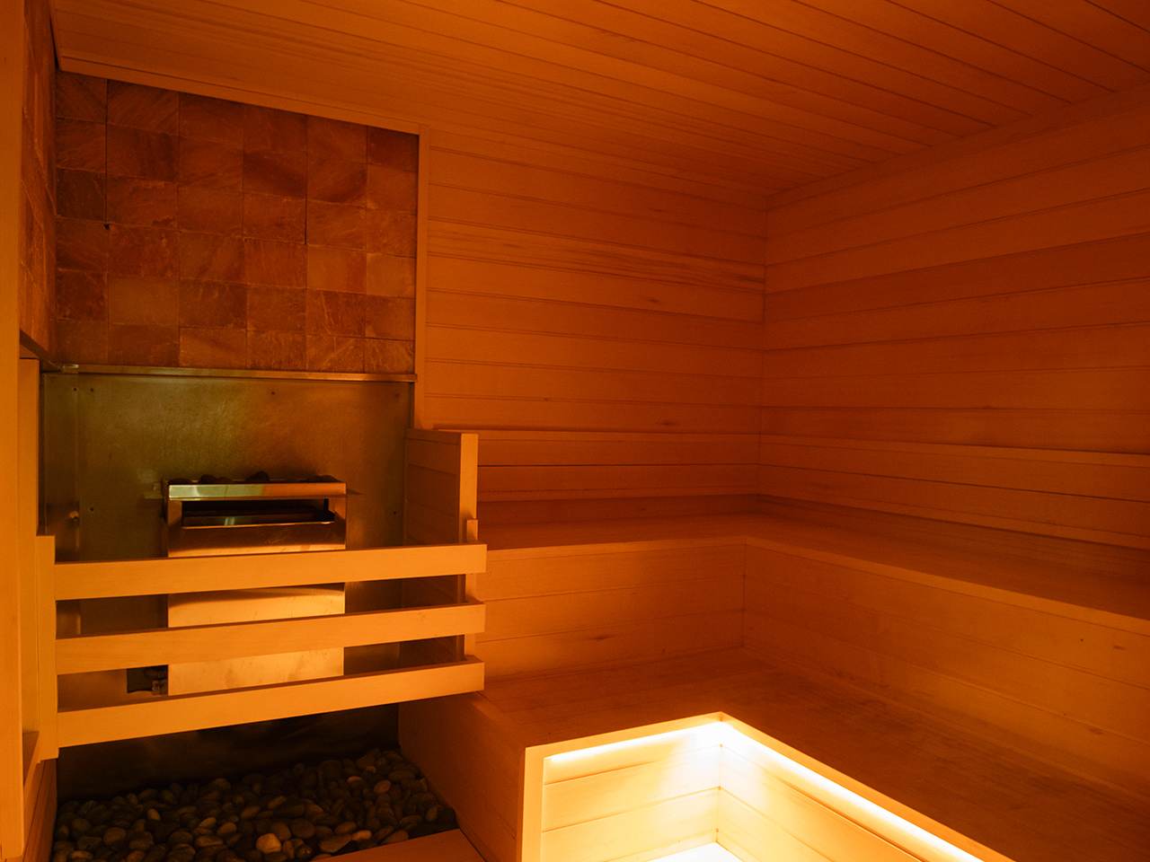 Wood-paneled sauna room with a stone heater on the left and built-in, illuminated seating along two walls—the ultimate recovery destination favored by personal trainers for relaxation and rejuvenation.