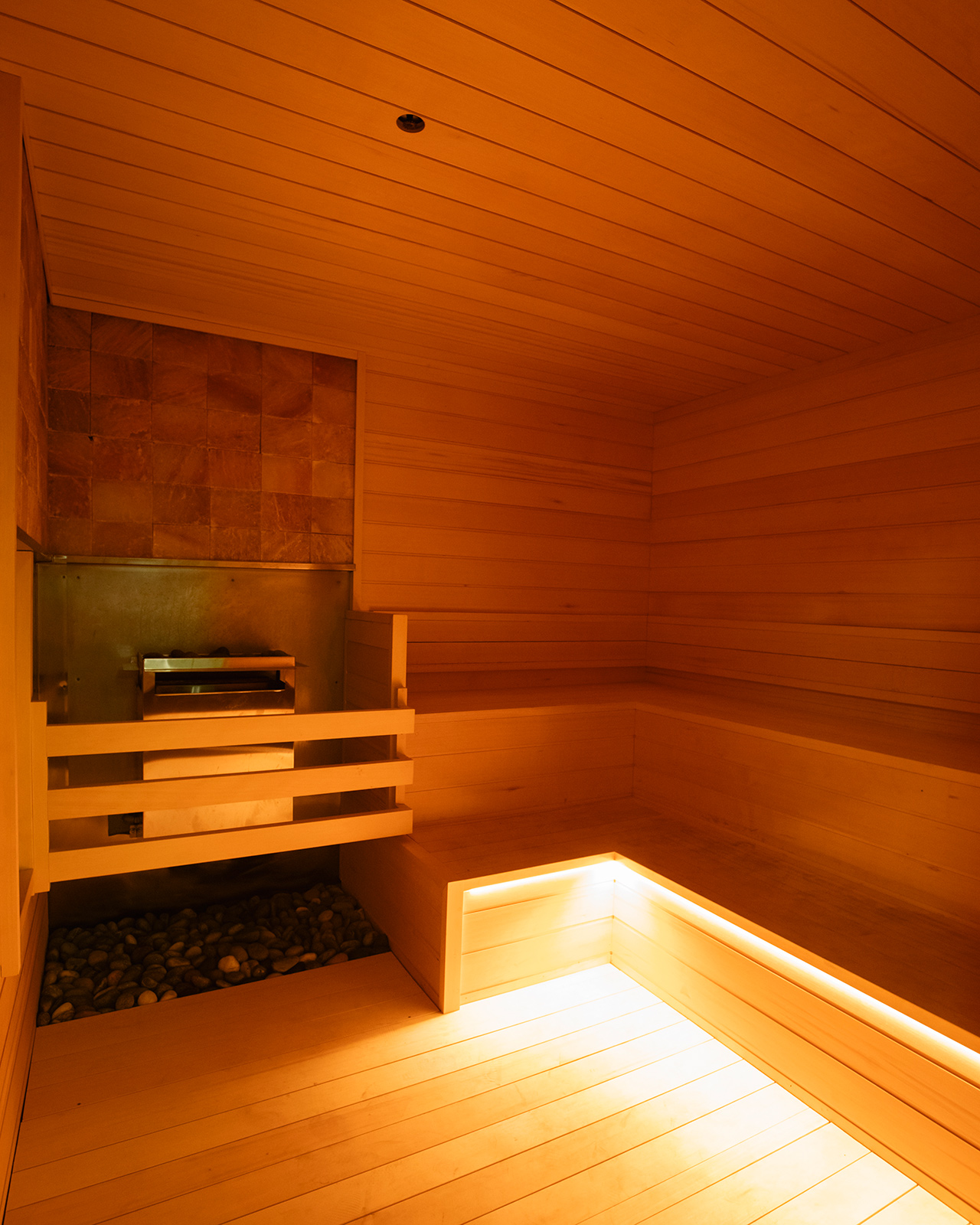 Wood-paneled sauna room with a stone heater on the left and built-in, illuminated seating along two walls—the ultimate recovery destination favored by personal trainers for relaxation and rejuvenation.