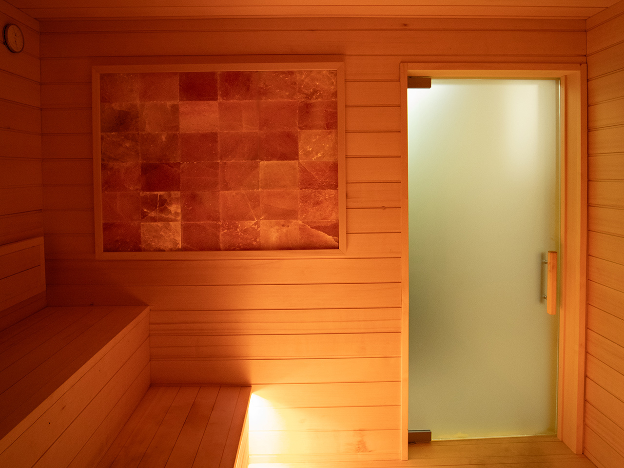 Interior of a wooden sauna with bench seating, a wall of salt bricks, and an open frosted glass door. Warm lighting creates a cozy atmosphere, making this the ultimate recovery destination—perfect for unwinding after sessions with personal trainers.