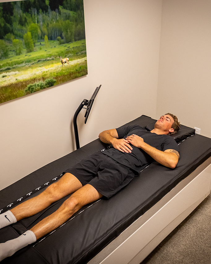 A man in athletic wear lies on his back on a therapy table in a small room with beige walls and a nature photo on the wall, receiving expert care from one of the facility’s personal trainers—an experience that defines this space as the ultimate recovery destination.
