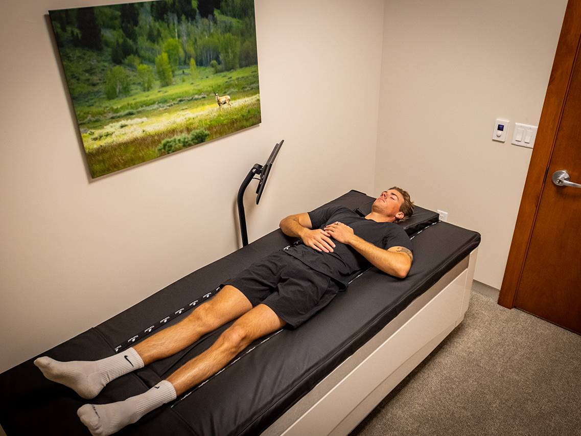 A man in athletic wear lies on his back on a therapy bed in a small room with a landscape photo on the wall and a closed wooden door, showcasing the Ultimate Recovery Destination trusted by personal trainers.
