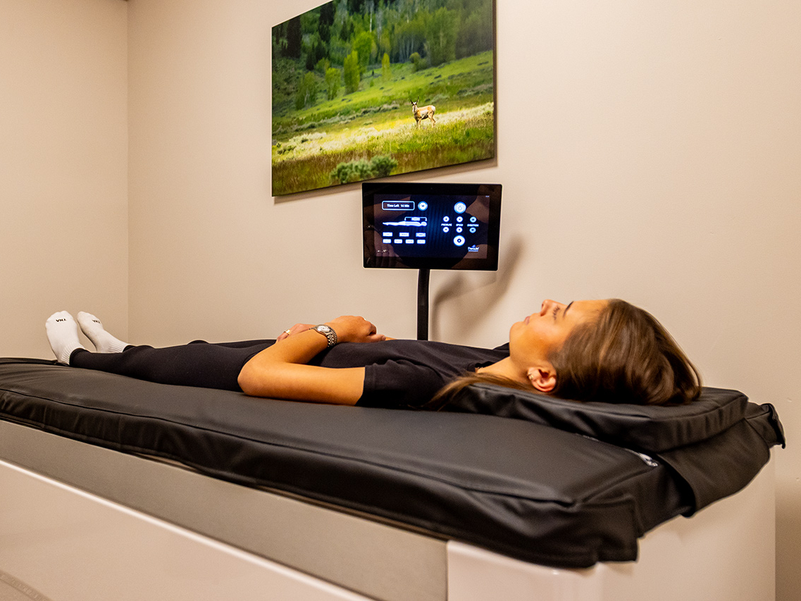 A person lies on a medical scanning table under a wall-mounted screen displaying controls, with a nature photograph on the wall above—an atmosphere reminiscent of the Ultimate Recovery Destination, where expert personal trainers guide every step of your wellness journey.