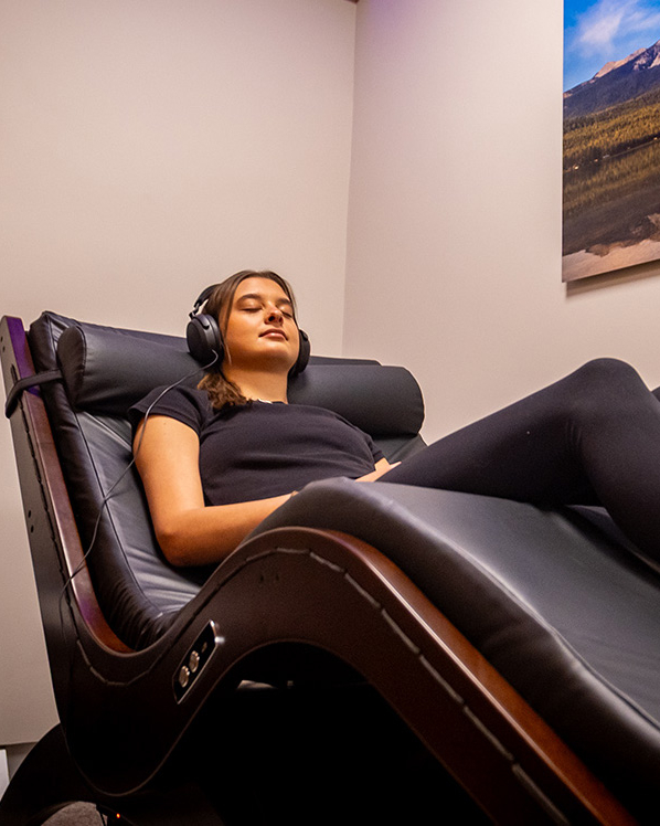 A person wearing headphones relaxes on a black recliner chair in a softly lit room with a landscape photo on the wall, creating the ultimate recovery destination after a session with personal trainers.
