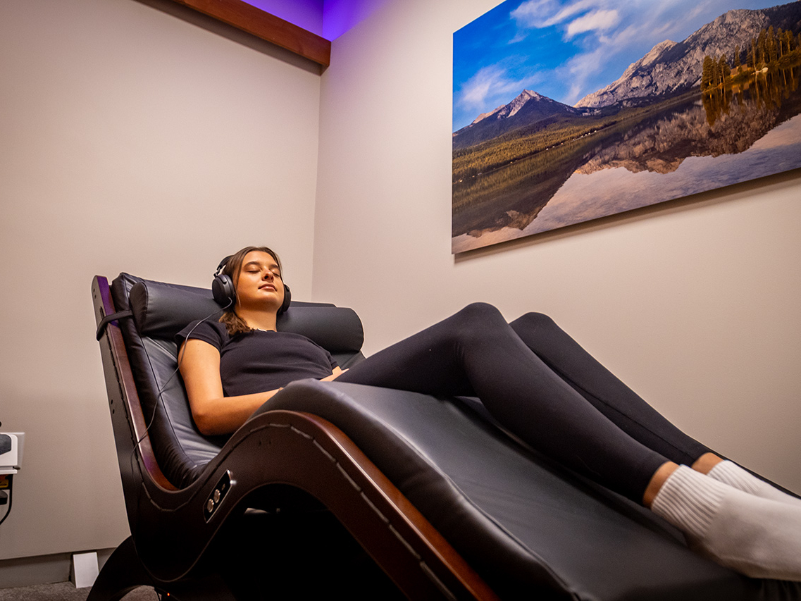 A person reclines on a black lounge chair wearing headphones in a softly lit room with a large landscape photo on the wall, creating the ultimate recovery destination where even personal trainers come to unwind.