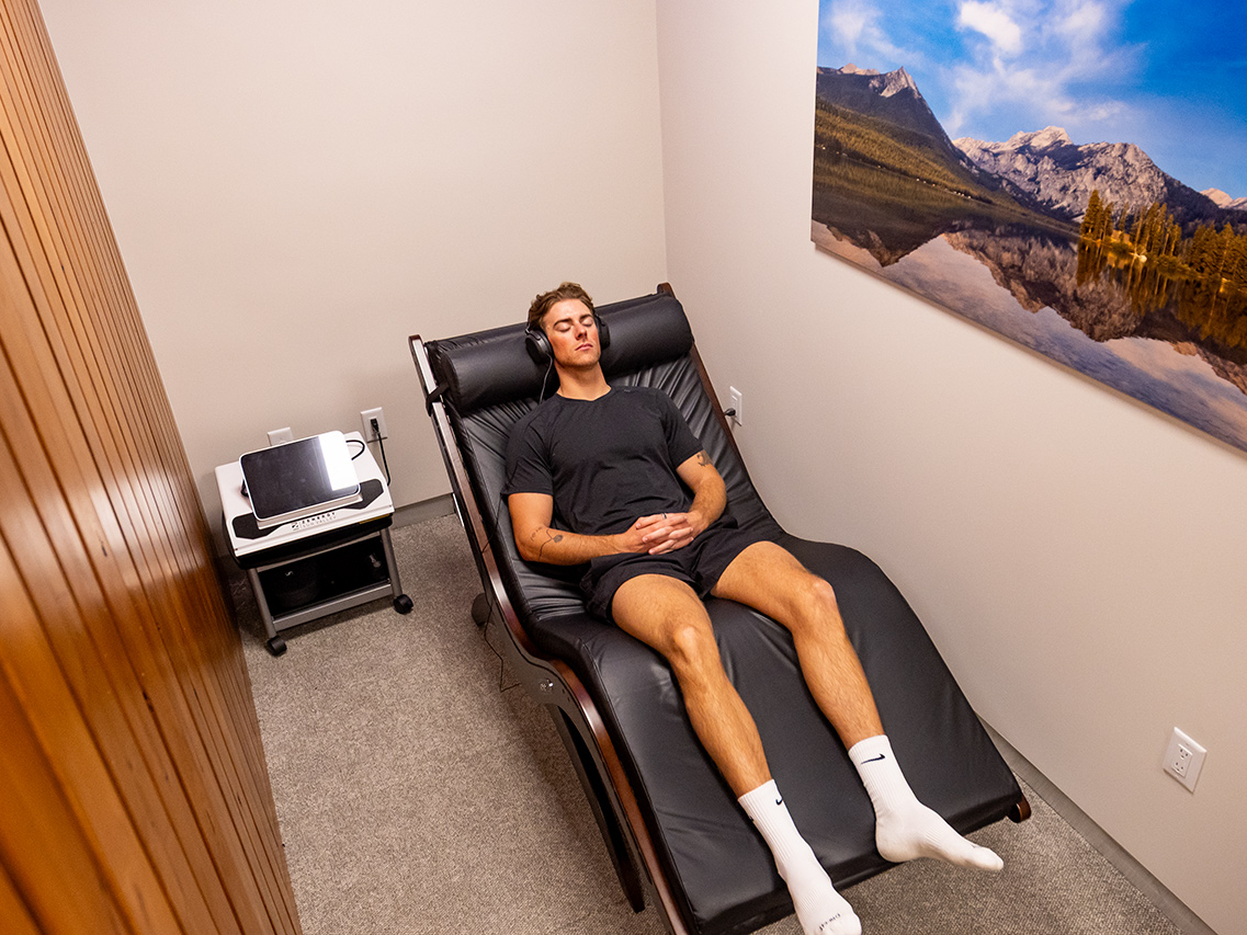 A man in athletic wear relaxes on a reclining chair in a small room with beige walls and a landscape photo hanging on the wall, making it the ultimate recovery destination often recommended by personal trainers.