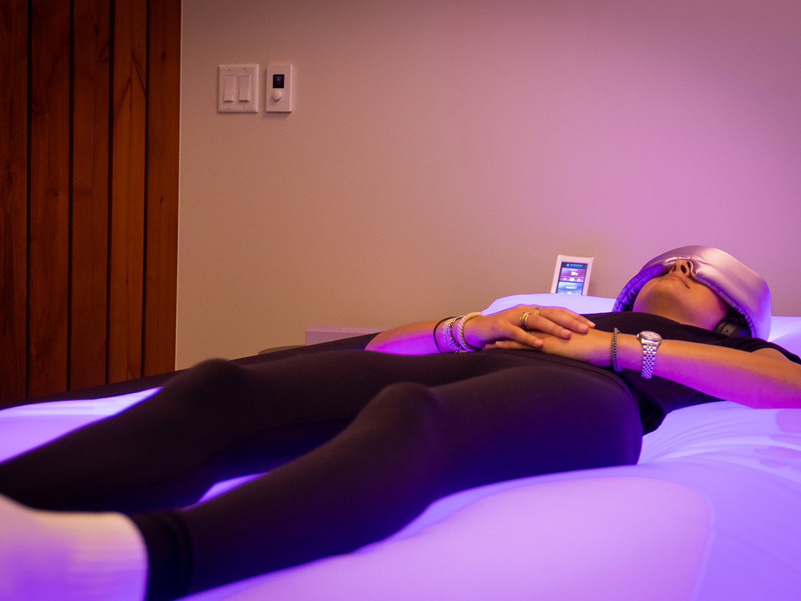Person lying on a glowing bed with a sleep mask, hands folded, in a dimly lit room with a control panel nearby—an ultimate recovery destination designed for complete relaxation and rejuvenation, perfect even for personal trainers seeking peak restoration.
