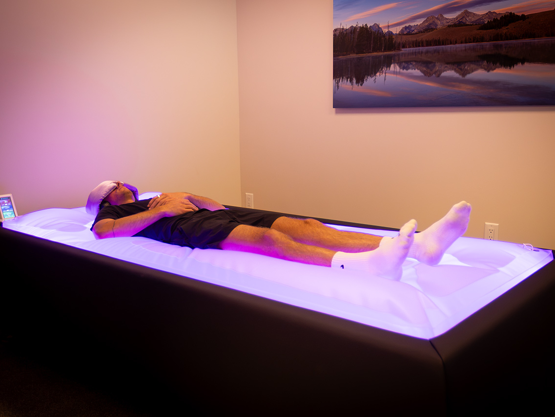A person wearing an eye mask and socks lies on a glowing water massage bed in a dimly lit room with a landscape photo on the wall, enjoying the ultimate recovery destination favored by personal trainers.