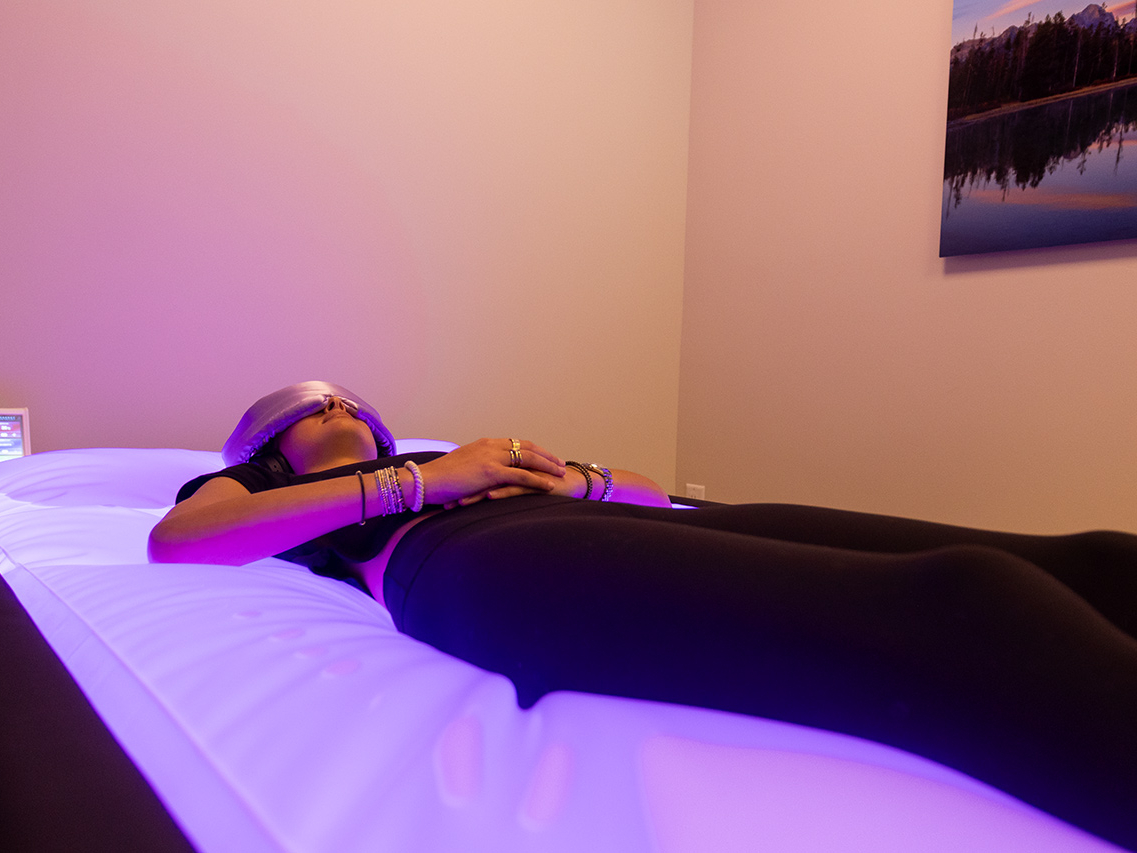 A person lies on a glowing massage bed with hands resting on their torso, wearing an eye mask in a dimly lit room with a landscape photo on the wall—an atmosphere that truly feels like the ultimate recovery destination.