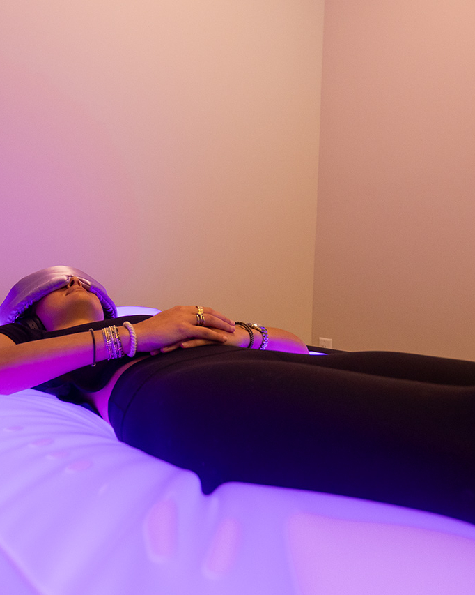 A person lies on a glowing purple spa bed with an eye mask, hands resting on their stomach, in a softly lit room with a wall-mounted landscape photo—an ultimate recovery destination often recommended by personal trainers.